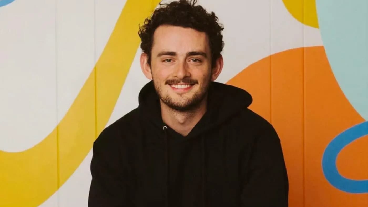 A smiling individual wearing a black hoodie sits in front of a colorful background featuring abstract shapes in yellow, orange, and blue.